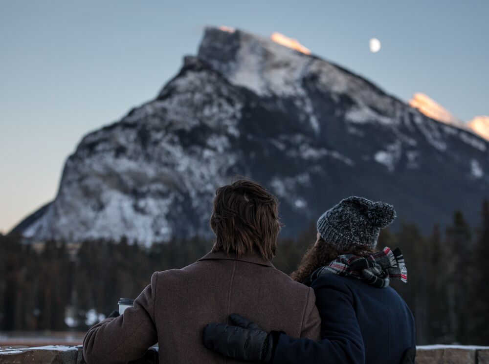 Two people look over the river towards Mount Rundle in Banff National Park at sunset.