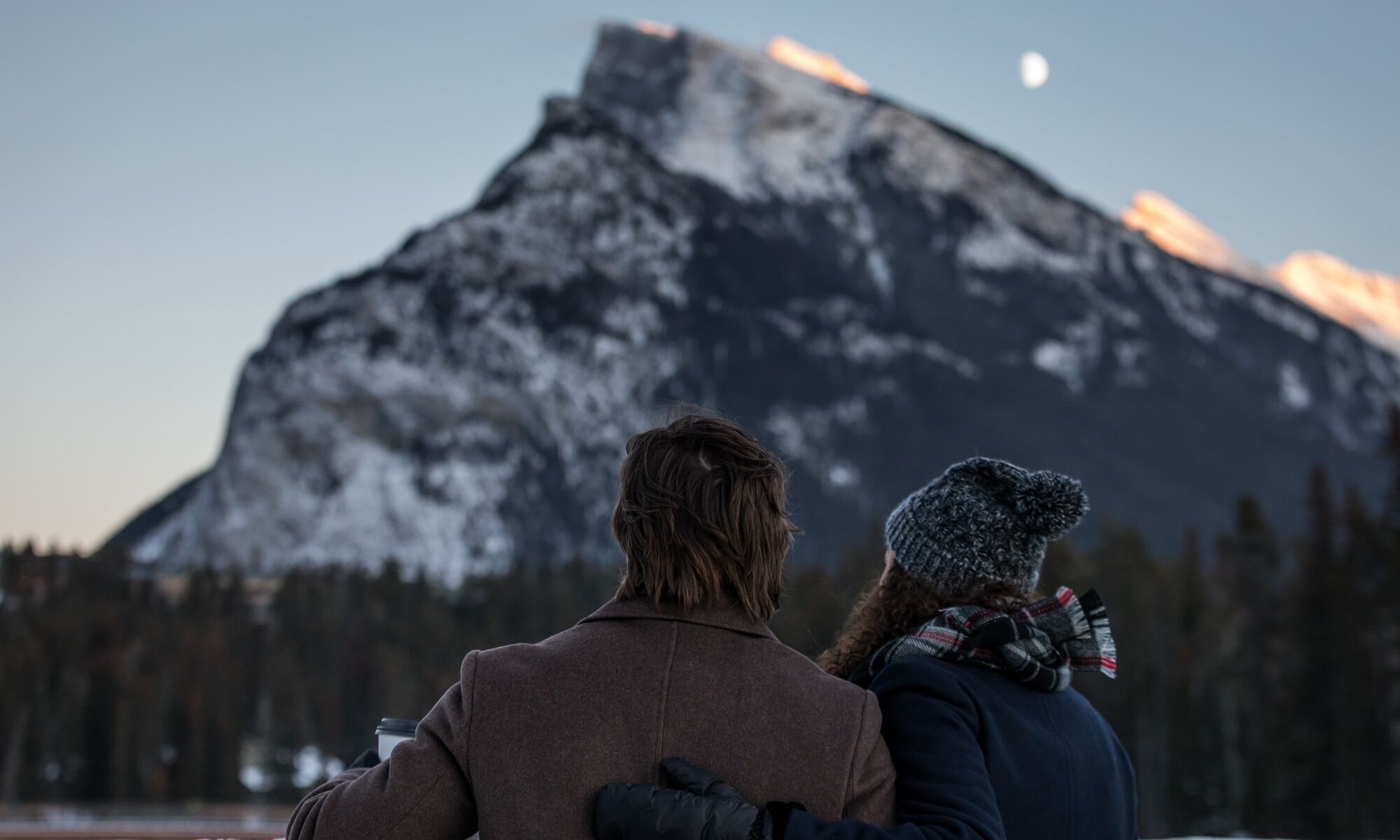 Two people look over the river towards Mount Rundle in Banff National Park at sunset.