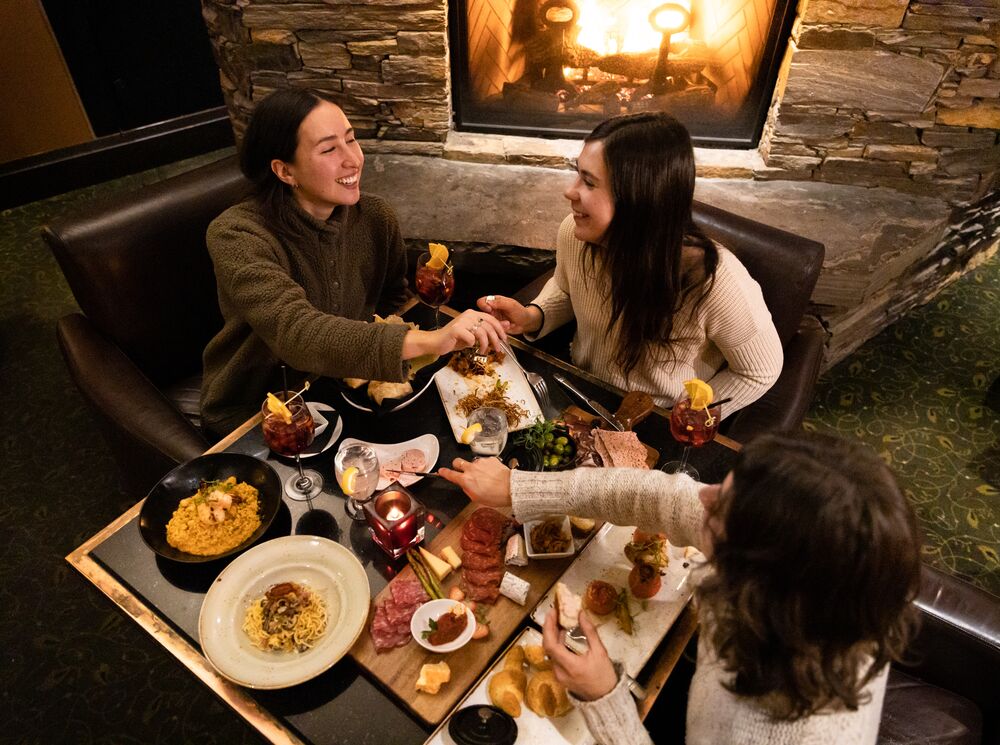 Three ladies enjoy a meal in Banff National Park. There are cheeses, meats, bread and wine on the table as seen from above and a fireplace behind them.