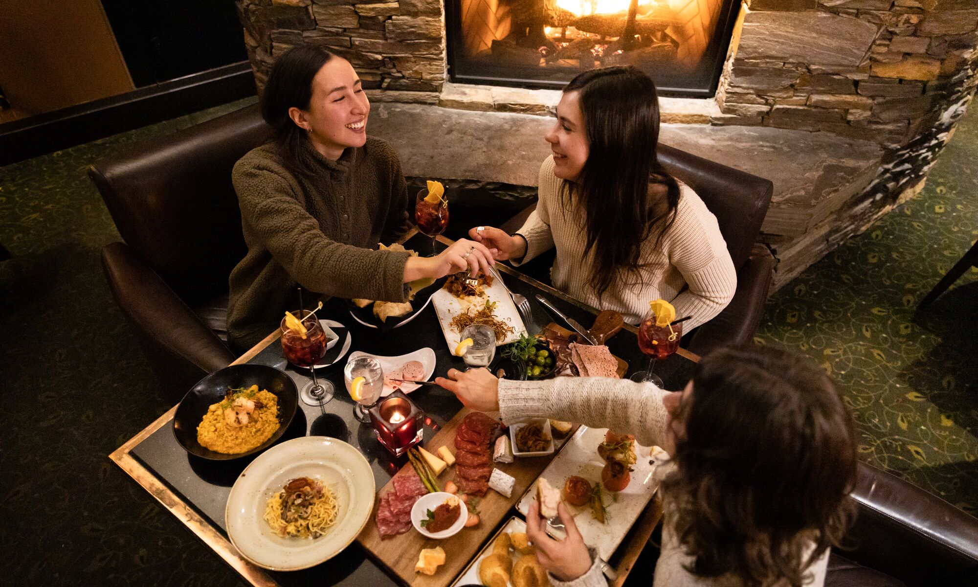 Three ladies enjoy a meal in Banff National Park. There are cheeses, meats, bread and wine on the table as seen from above and a fireplace behind them.