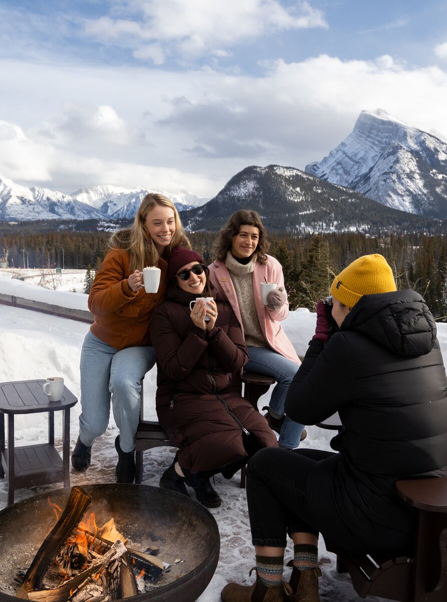 Four women on the outside patio in winter at Juniper Bistro enjoying warm drinks with Mount Rundle and Tunnel Mountain behind them in Banff National Park.