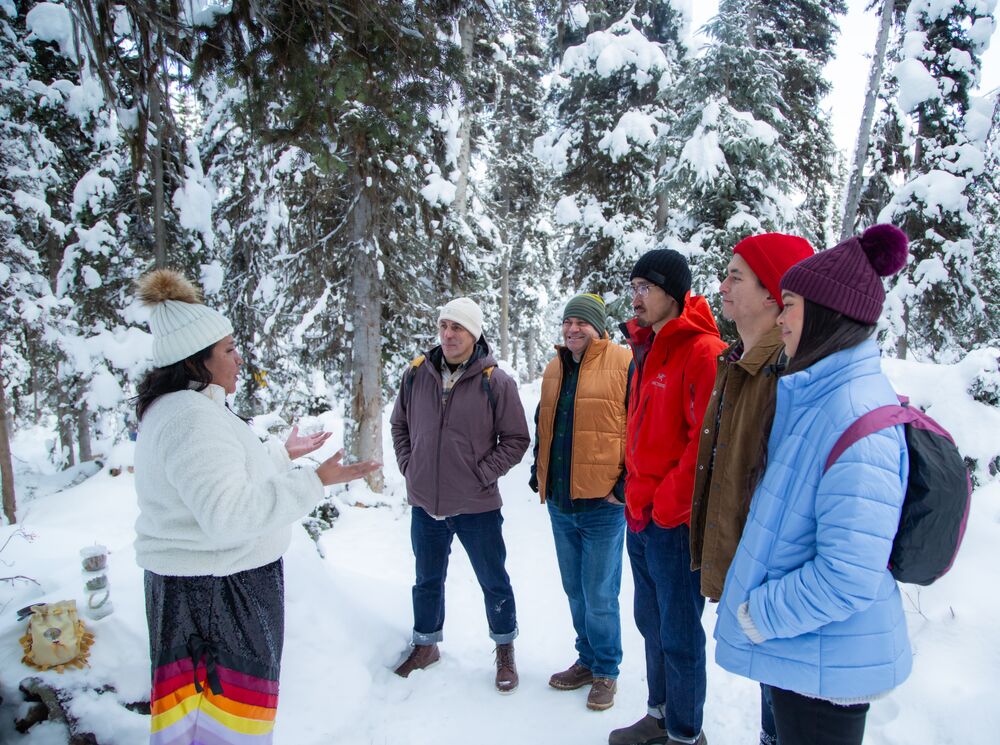 A group of people standing around a women listening to her speak in a snow covered forest.