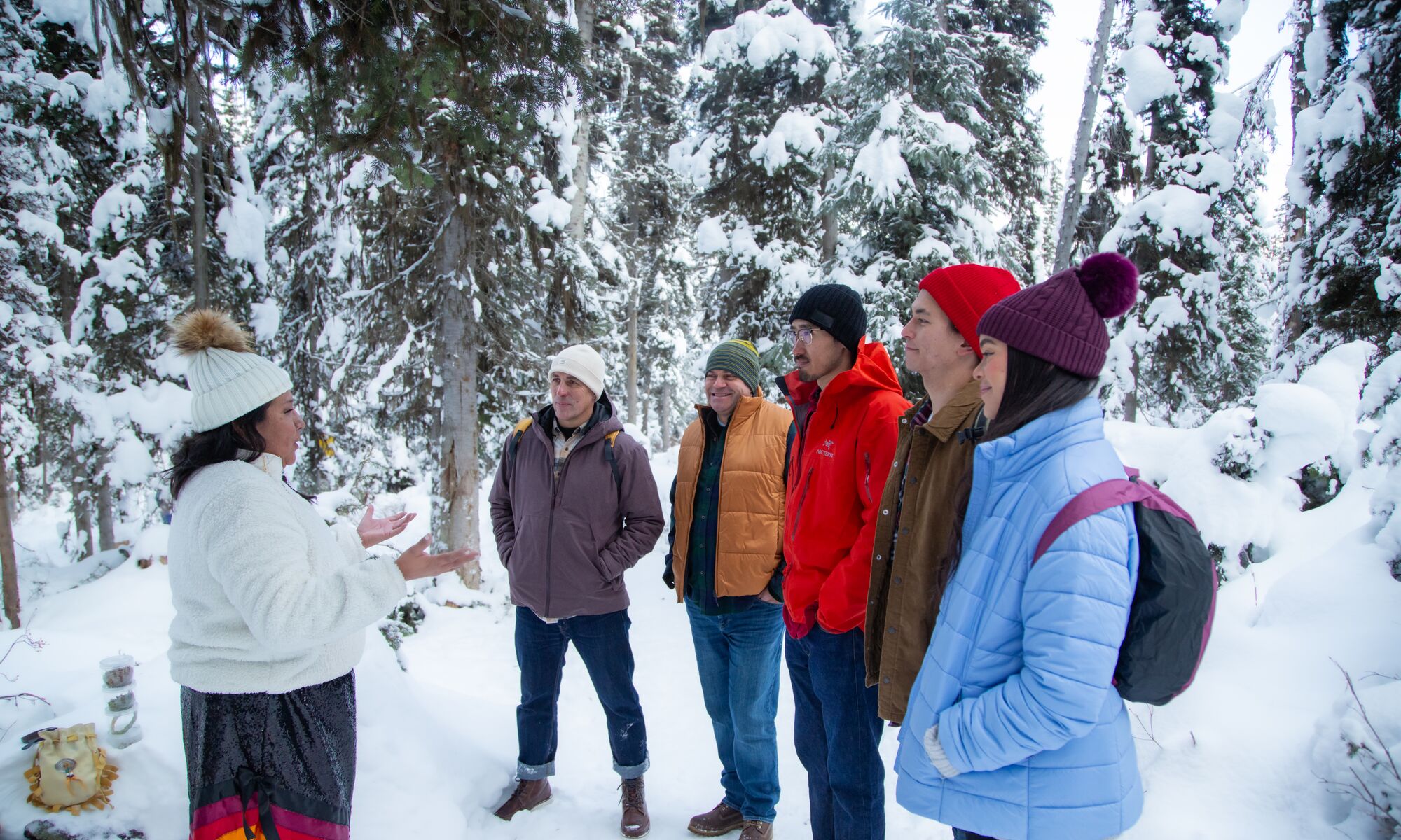 A group of people standing around a women listening to her speak in a snow covered forest.