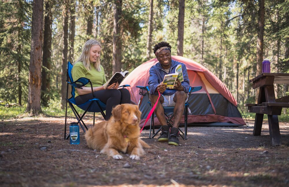 Two people sitting on camp chairs with a dog in front of them and their camp set up in the background