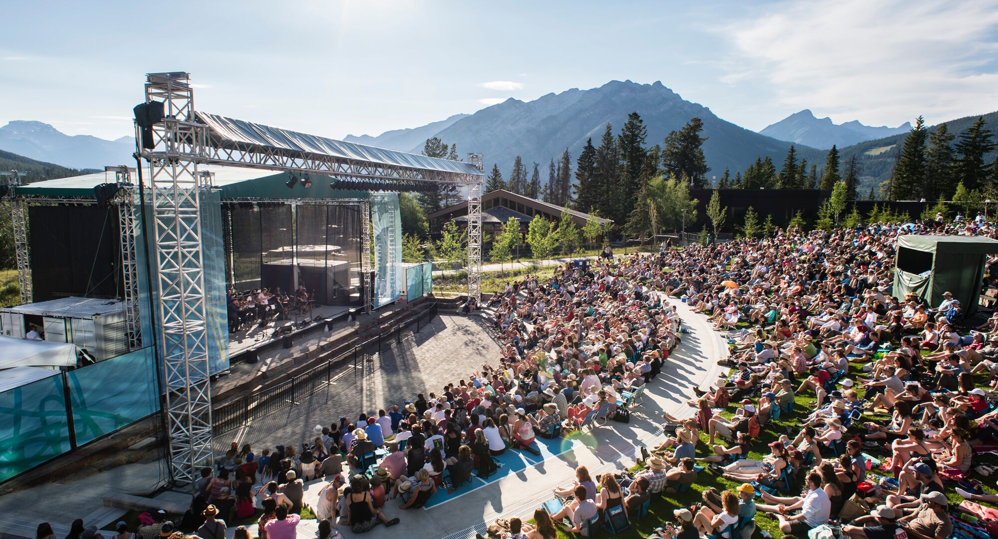 A large crowd is seated in a semicircle around an outdoor stage while watching a performance on a sunny day at the Banff Centre