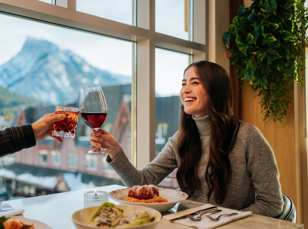 A woman clinks her wine glass with another glass while enjoying the mountain views out of the third floor window at LUPO Italian Ristorante in Banff, Canada.