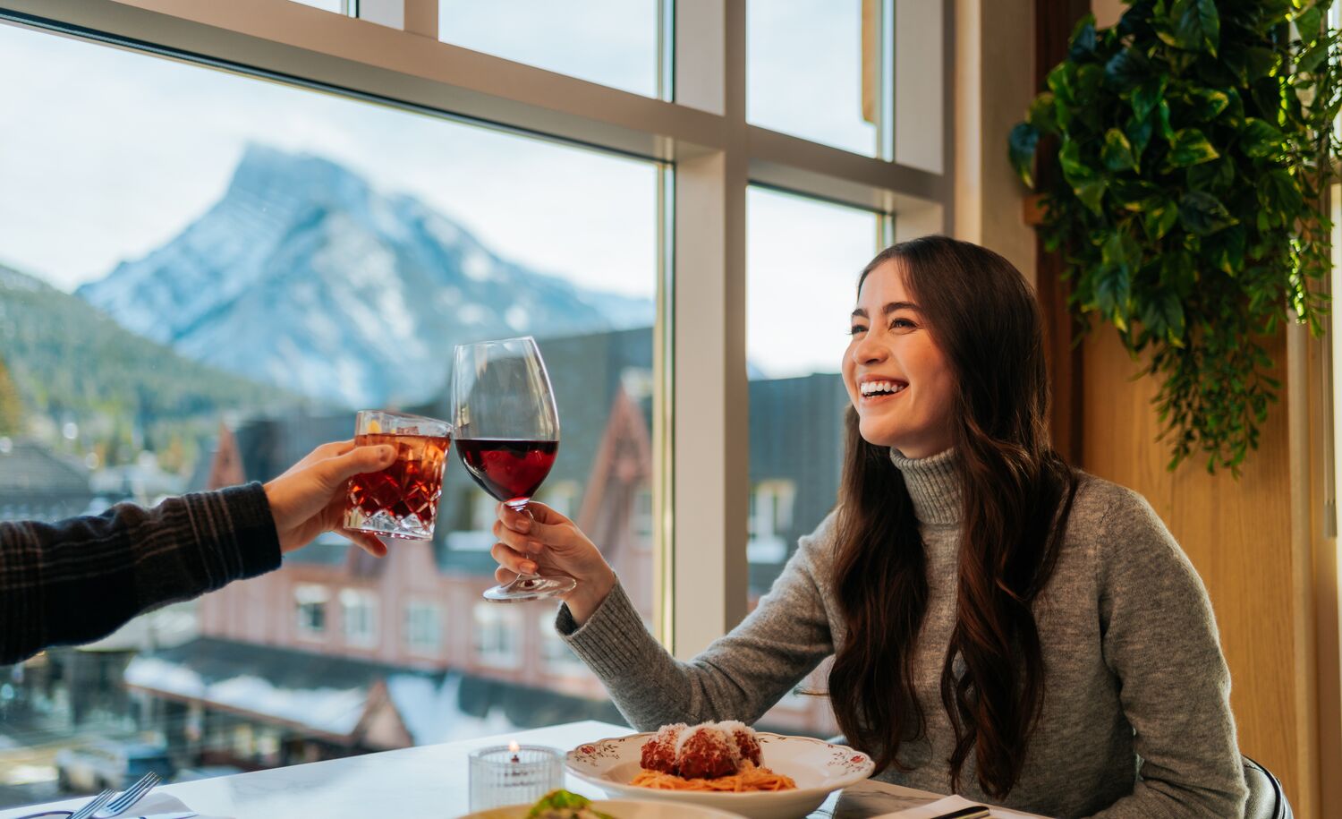 A woman clinks her wine glass with another glass while enjoying the mountain views out of the third floor window at LUPO Italian Ristorante in Banff, Canada.