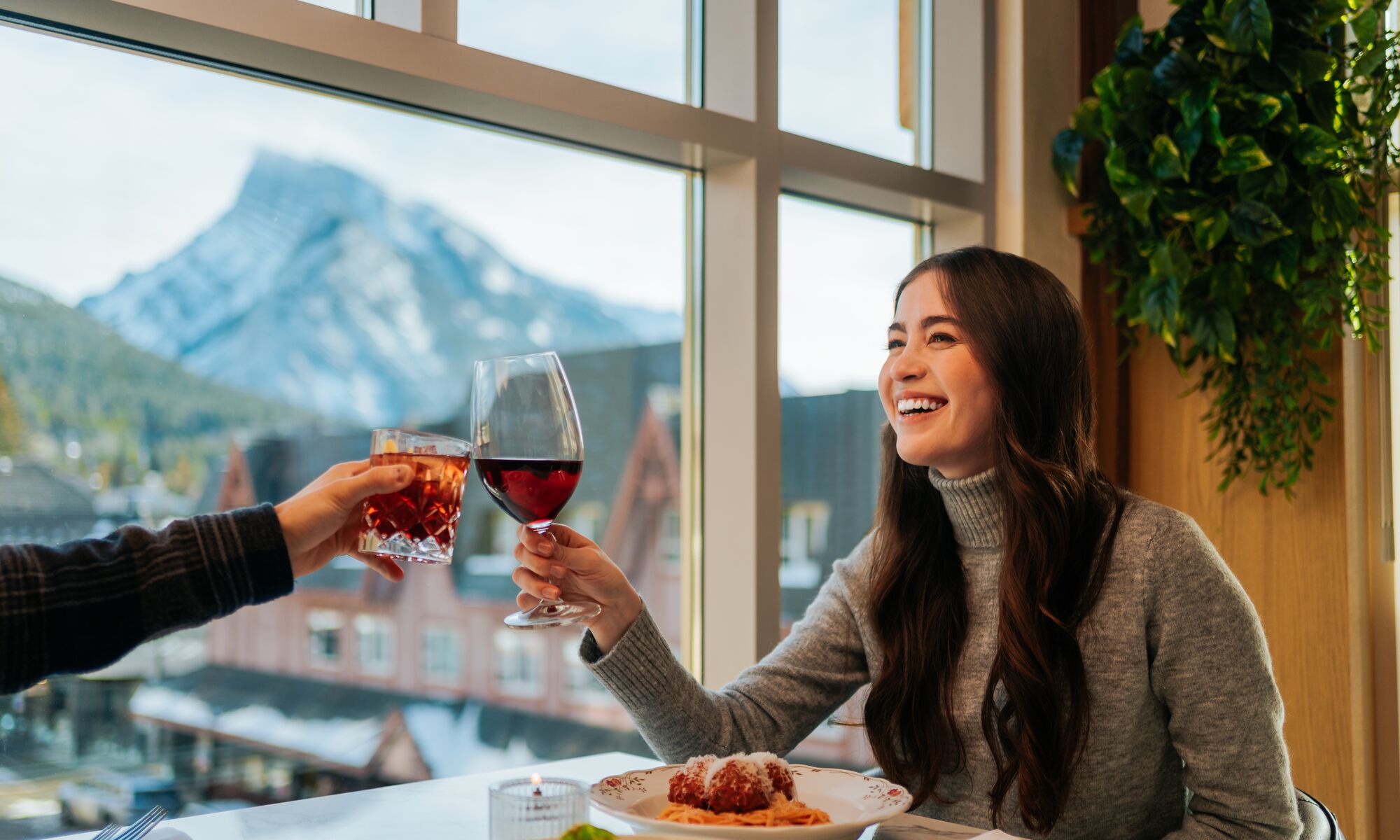 A woman clinks her wine glass with another glass while enjoying the mountain views out of the third floor window at LUPO Italian Ristorante in Banff, Canada.