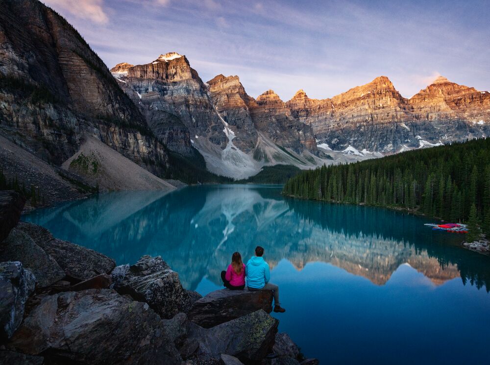 Two people sit watching sunrise at Moraine Lake with orange hues touching the mountain tops in Banff National Park.