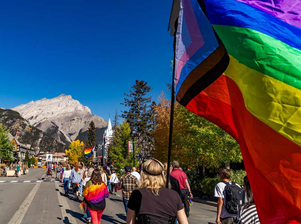 A Progressive Pride Flag flies close to the camera during the Banff Pride March on Banff Ave with Cascade Mountain in the background.