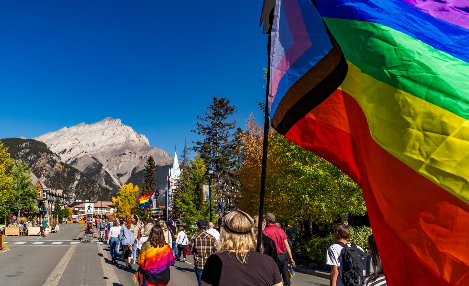 A Progressive Pride Flag flies close to the camera during the Banff Pride March on Banff Ave with Cascade Mountain in the background.