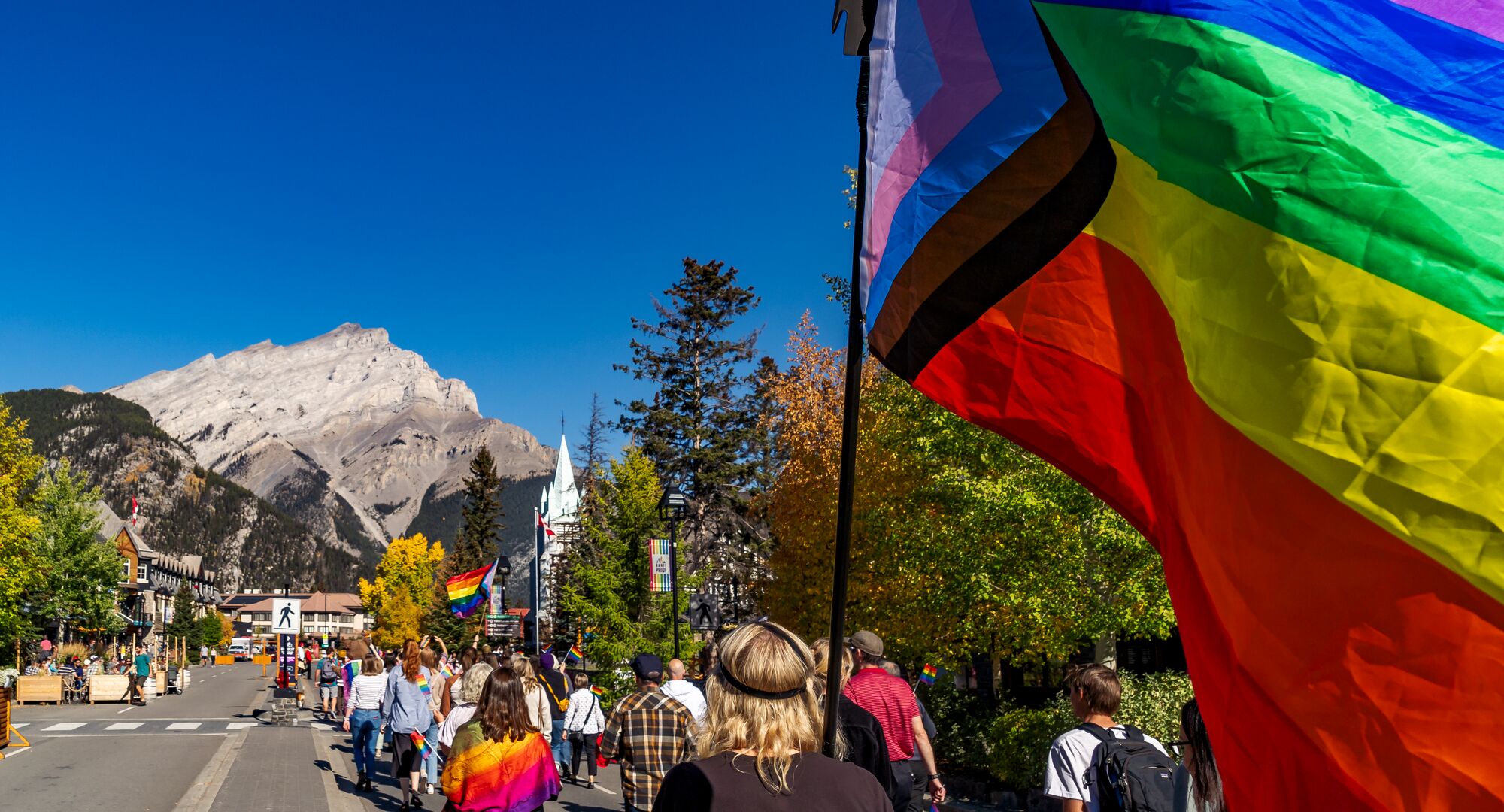 A pride flag in the Banff Pride parade on Banff Ave with Cascade Mountain in the background.