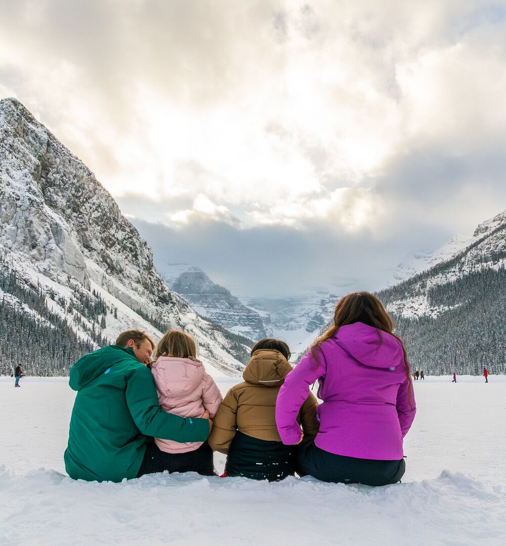 Family sitting on the shores of snowy Lake Louise looking at mountains in the winter.