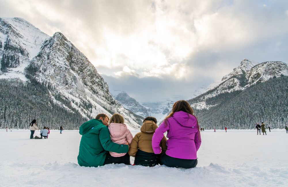Family sitting on the shores of snowy Lake Louise looking at mountains in the winter.