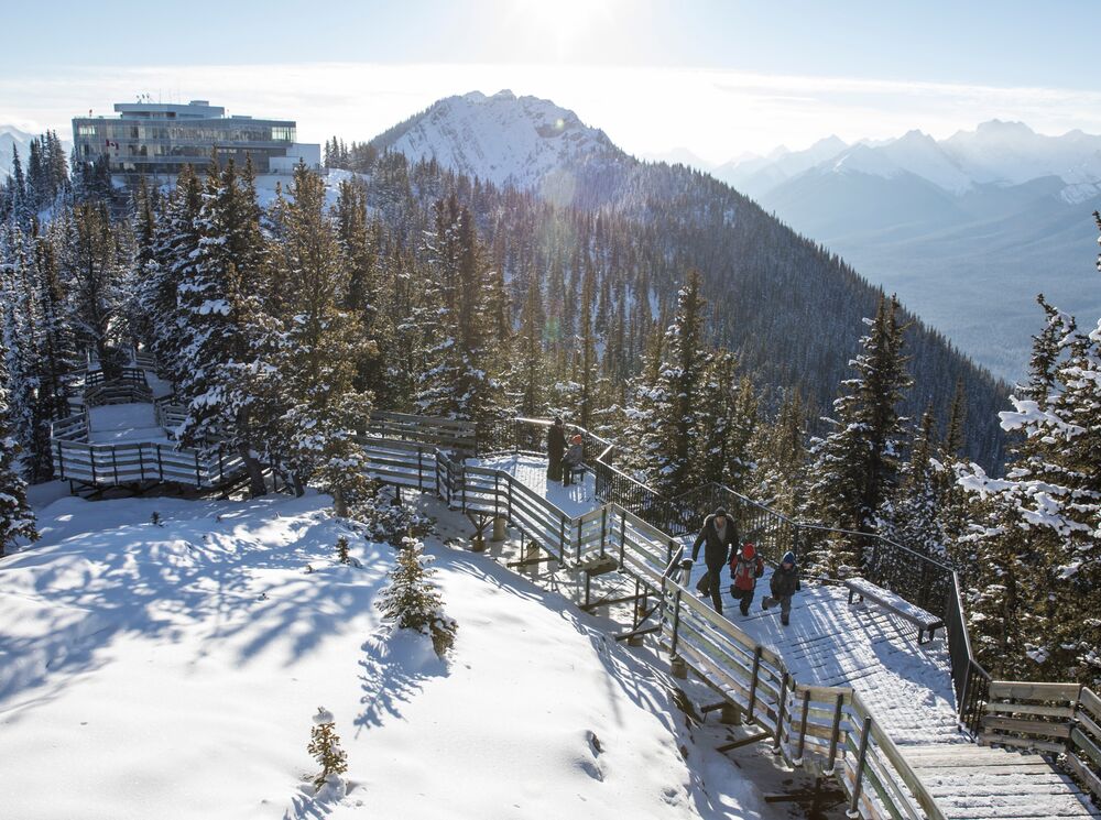 The family explores the summit boardwalk from the Banff Gondola, enjoying the views of snow-covered mountains surrounding them. 