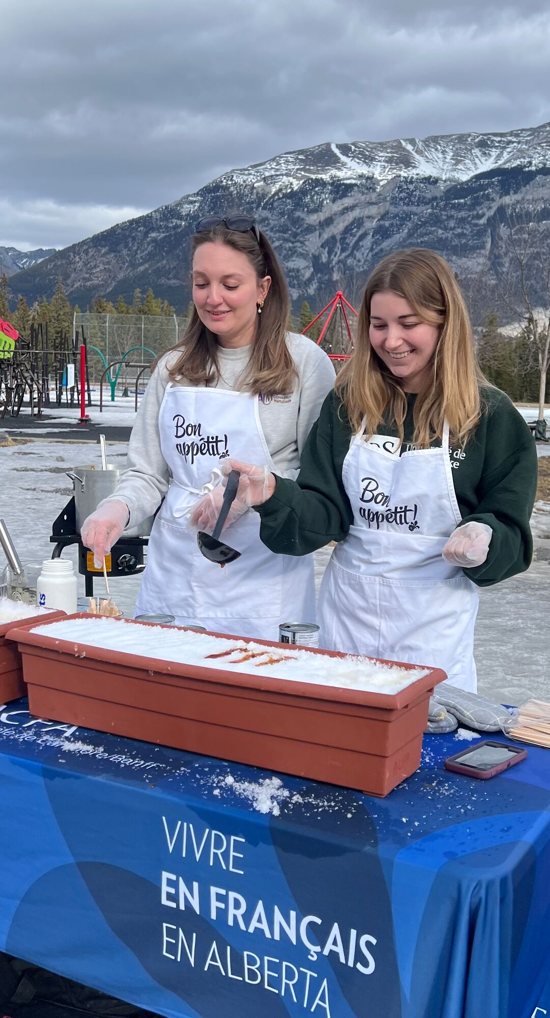 Two volunteers serving maple toffee poured over snow outdoors in Banff with mountain views, showcasing a traditional Sugar Shack winter activity.