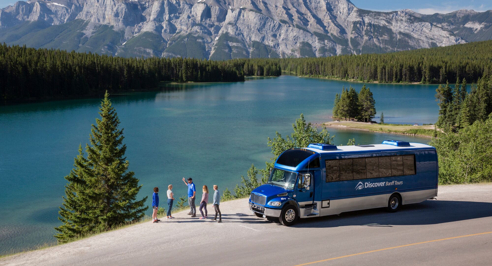 A tour group stop to admire a turquoise lake with Mount Rundle in the background. A Discover Banff Tours bus is parked nearby