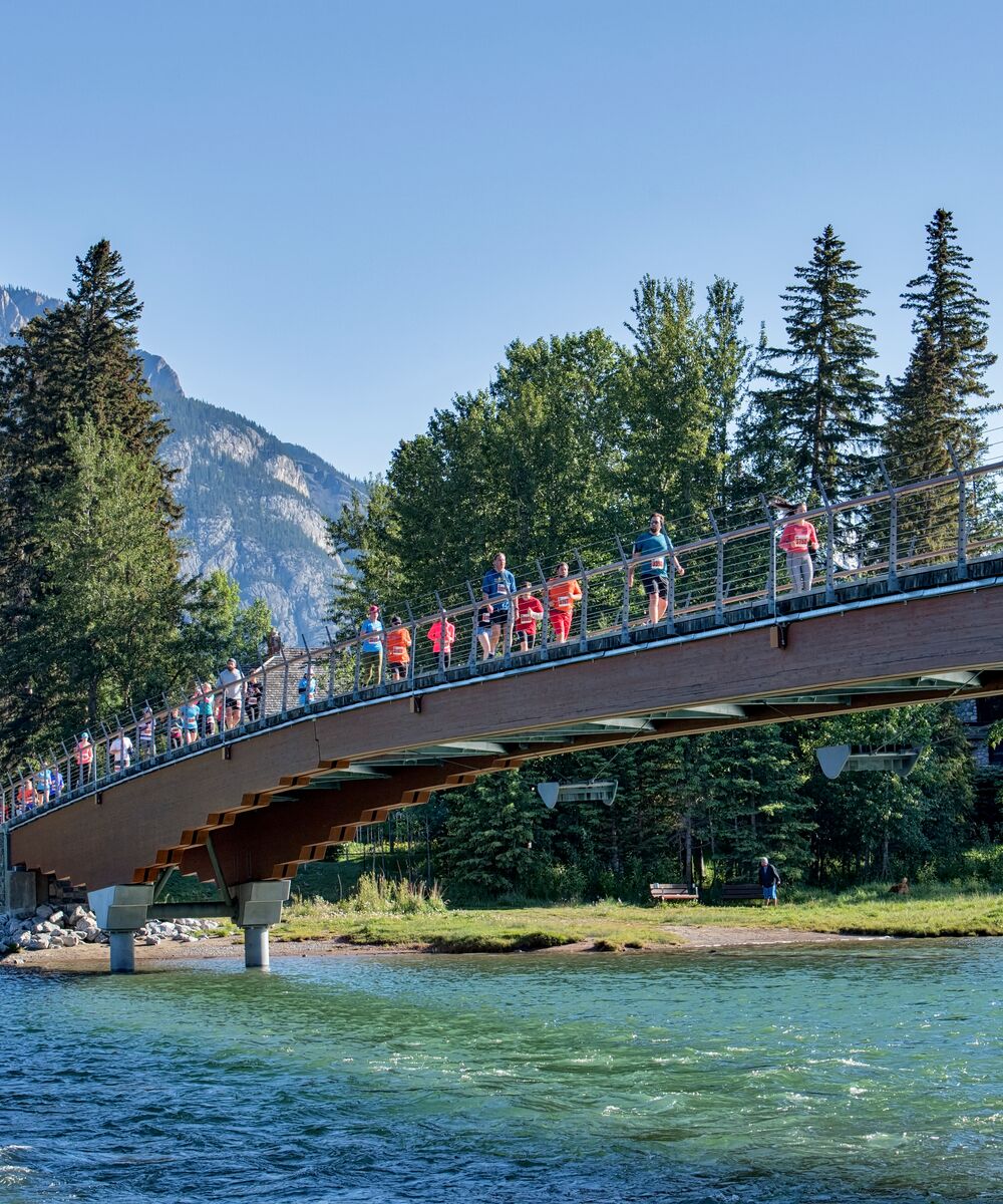 A group of runners participating in a marathon run across a pedestrian bridge over the Bow River with Cascade Mountain behind them on a clear summer day