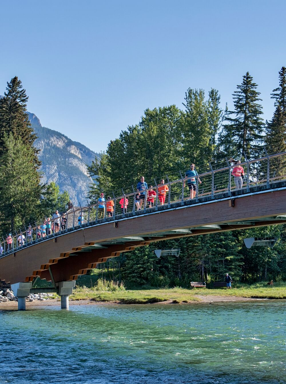 A group of runners participating in a marathon run across a pedestrian bridge over the Bow River with Cascade Mountain behind them on a clear summer day