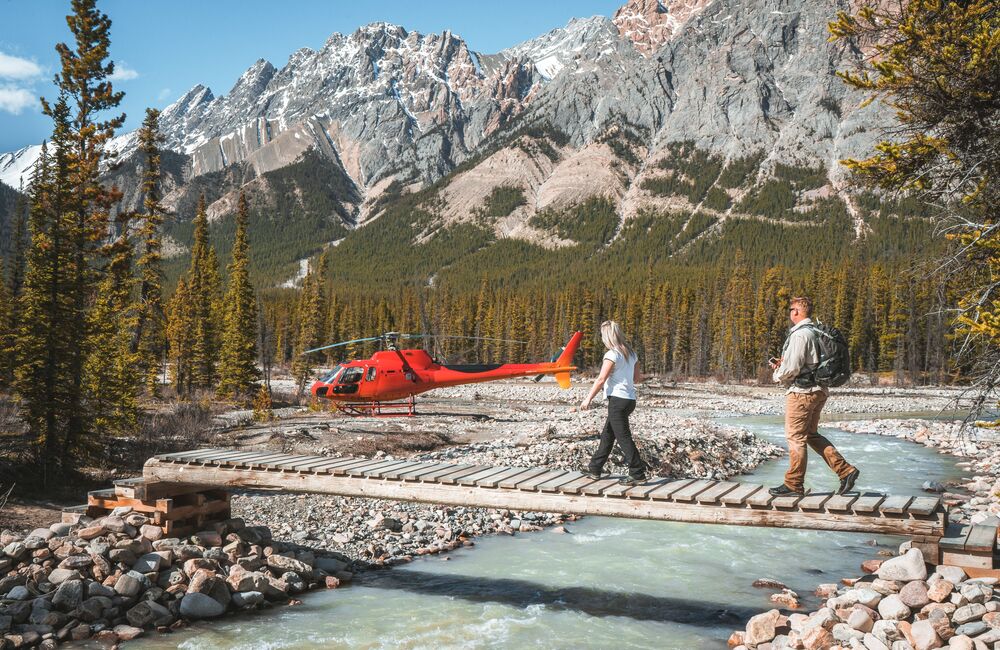 Two people hiking across a bridge towards a helicopter on a heli-hiking tour in the Canadian Rockies 