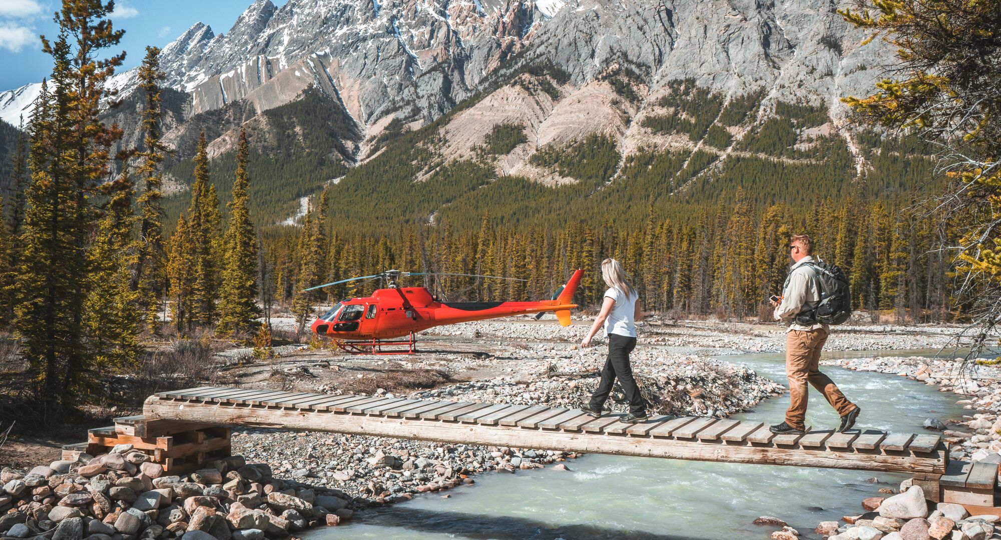 Two people crossing a bridge in front of a helicopter on a tour with Rockies Heli