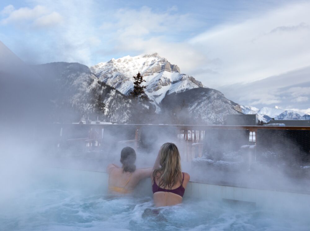 Two friends look at Cascade Mountain while enjoying a rooftop hot tub at the Moose Hotel in Banff.