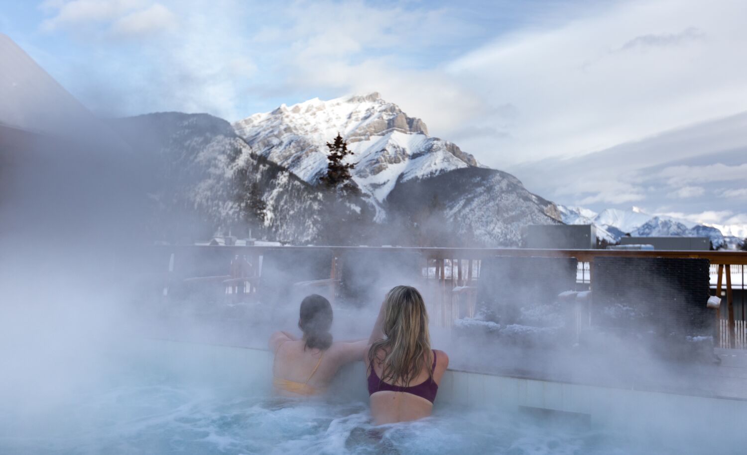 Two friends look at Cascade Mountain while enjoying a rooftop hot tub at the Moose Hotel in Banff.