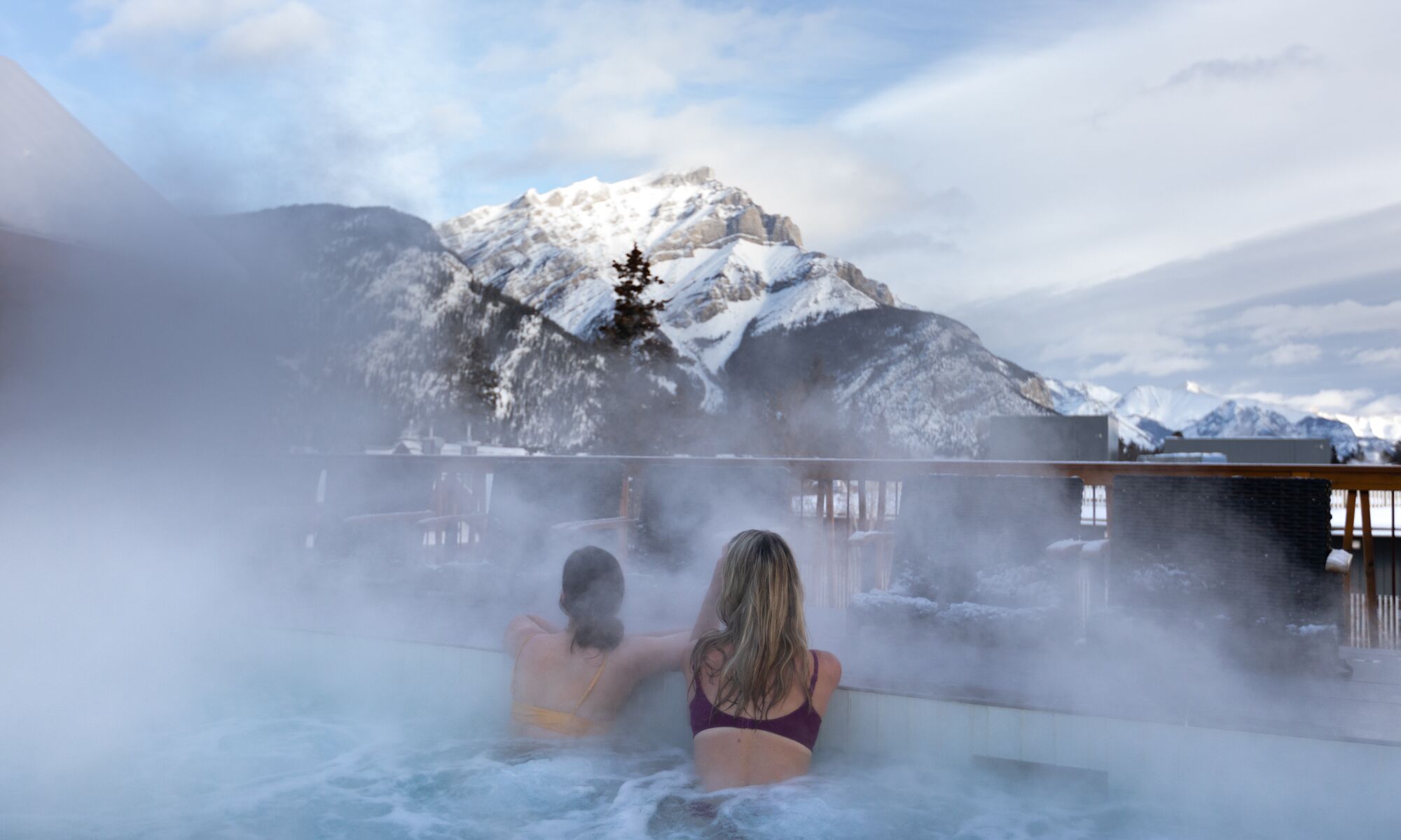Two friends look at Cascade Mountain while enjoying a rooftop hot tub at the Moose Hotel in Banff.