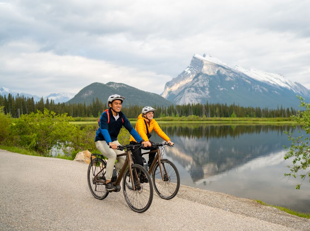 Two people biking Vermilion Lakes Drive with a view of Mount Rundle in the background