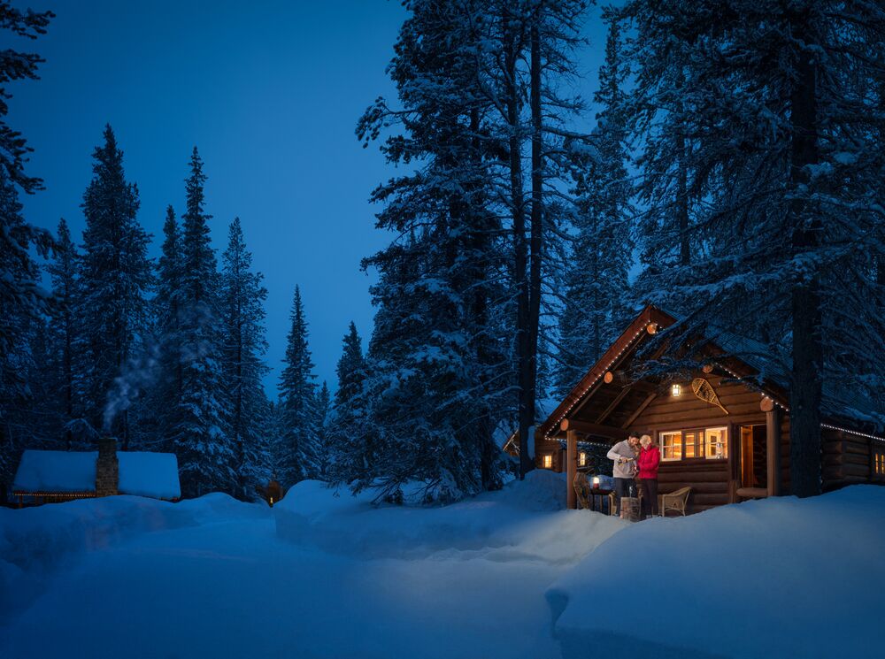 A couple stands at a log cabin in the snow at Storm Mountain Lodge in Banff National Park.