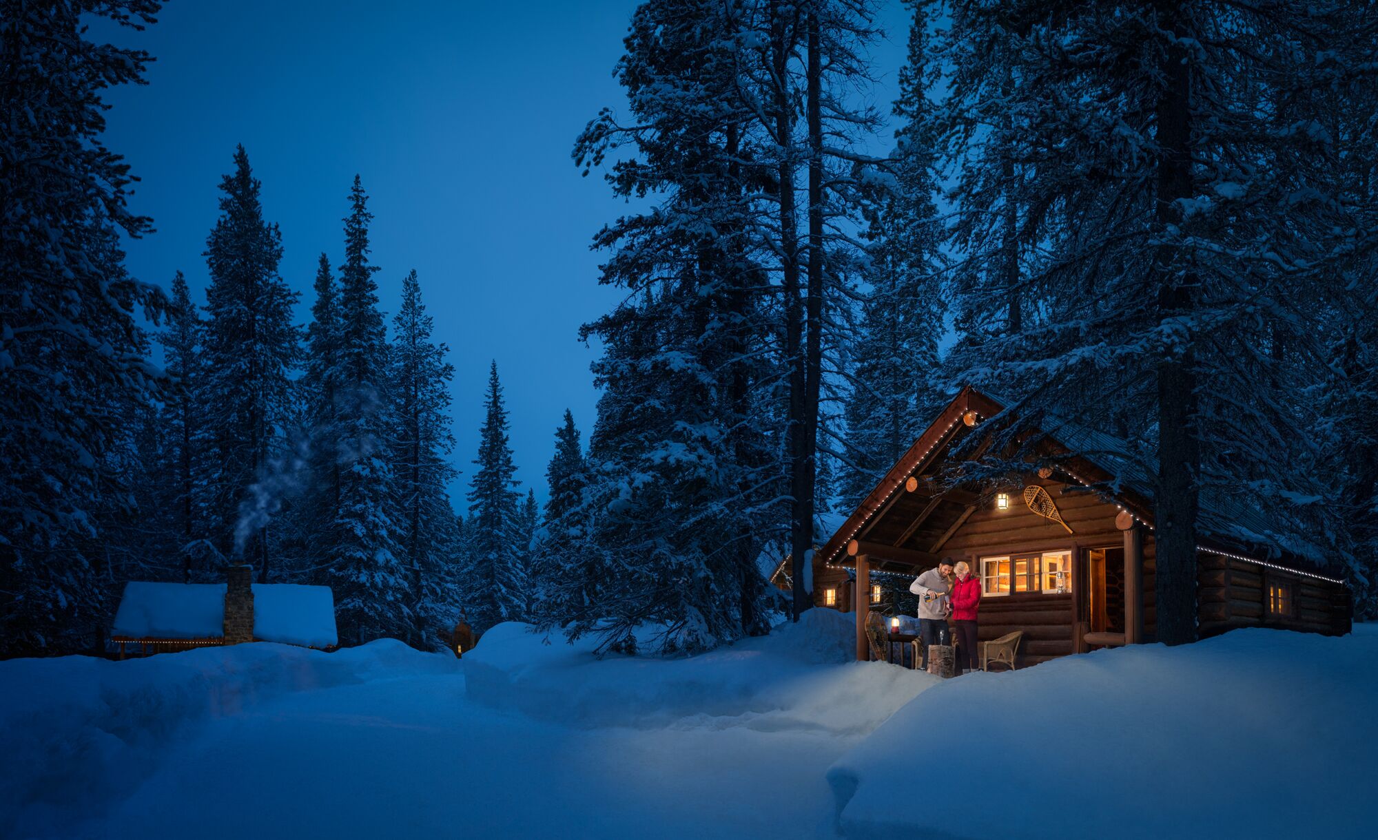 A couple stands at a log cabin in the snow at Storm Mountain Lodge in Banff National Park.