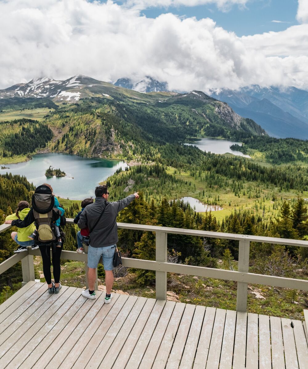 A family with two small children stand at a viewing deck overlooking alpine lakes and peaks in Sunshine Meadows