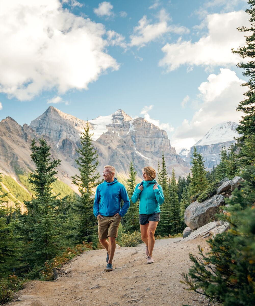 A couple hikes through a valley in Lake Louise with mountains surrounding them on a clear summer day
