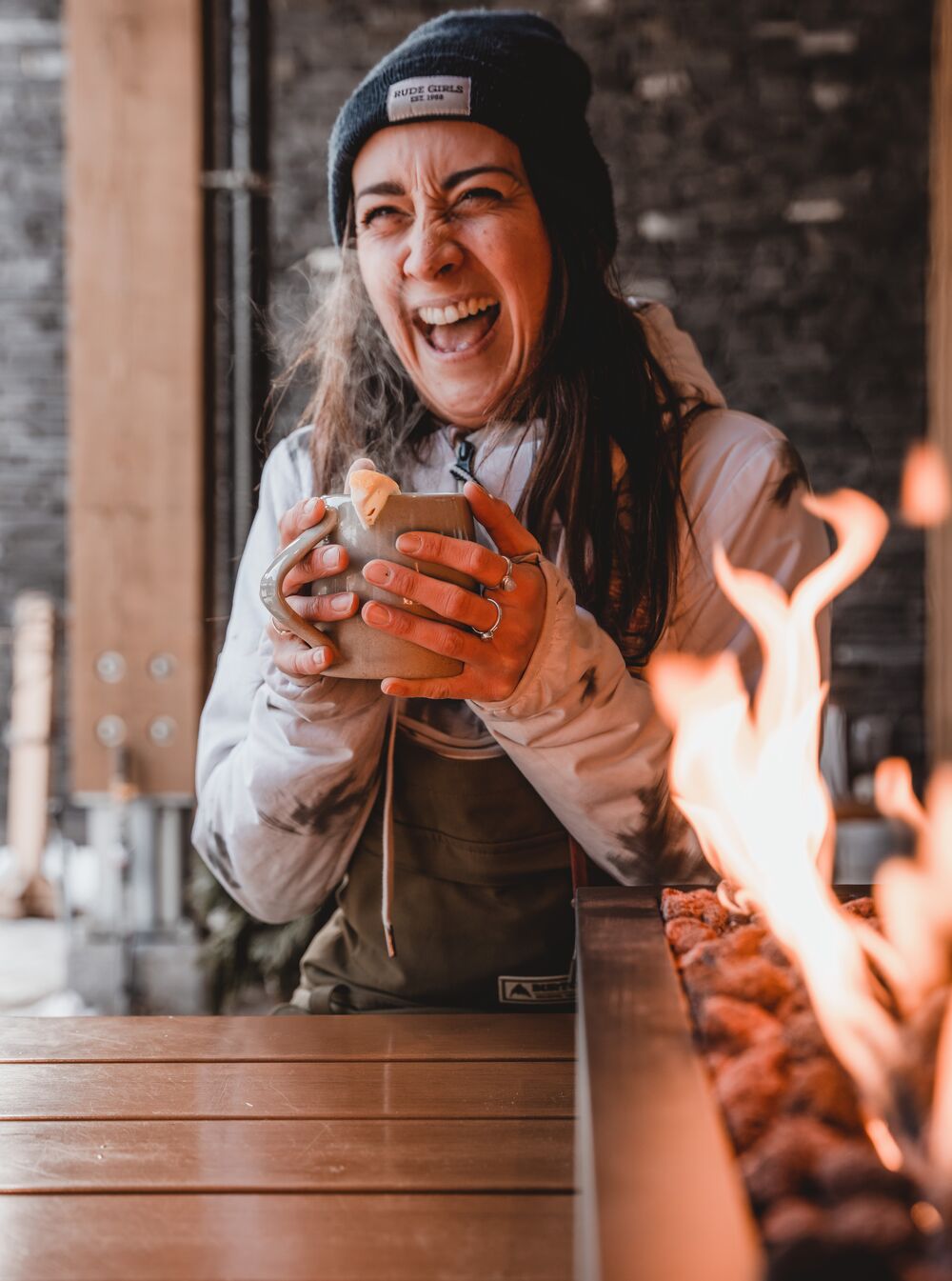 A woman smiles and laughs while wearing a toque and sitting near a table-fire at Three Bears Brewery in Banff National Park.