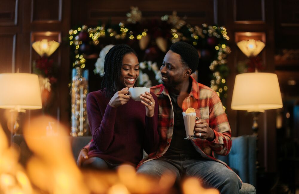 A couple enjoy hot chocolate sitting by a fire place with Christmas trees behind them at the Rimrock Hotel in Banff National Park.