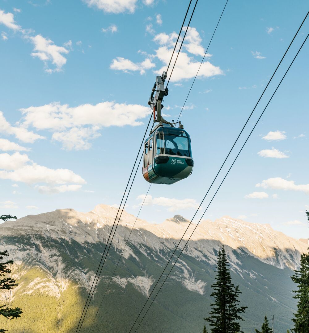 A gondola car goes up the cable with Mount Rundle in the background