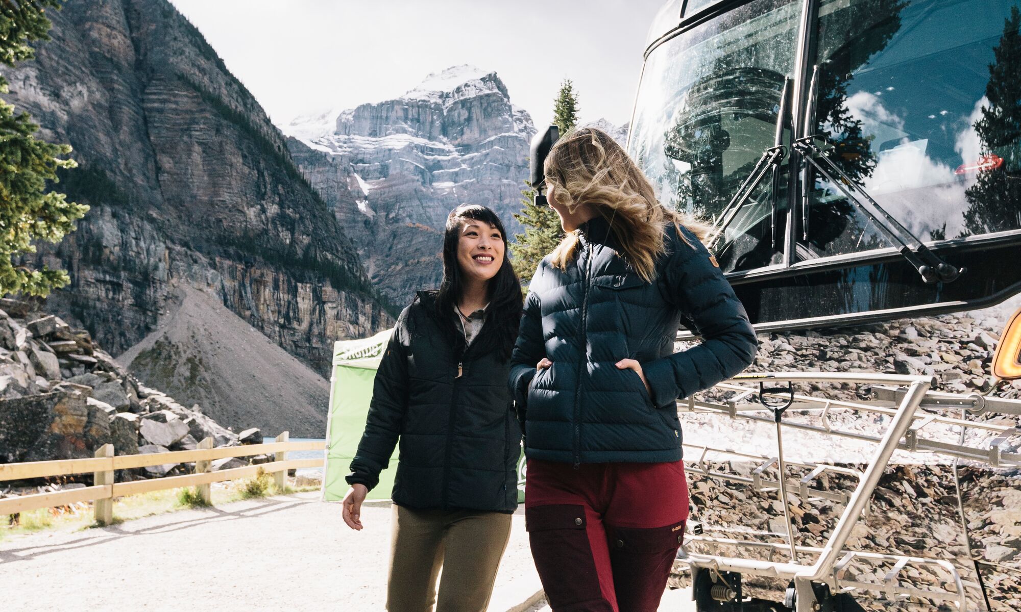 Two people in front of the Roam Public Transit bus at Moraine Lake on a sunny fall day in Banff National Park.