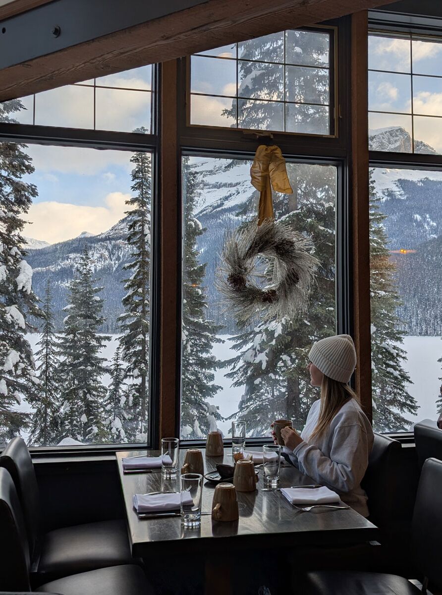 A woman sits at a restaurant table near large windows at Emerald Lake Lodge in Yoho, Canada near Banff National Park.