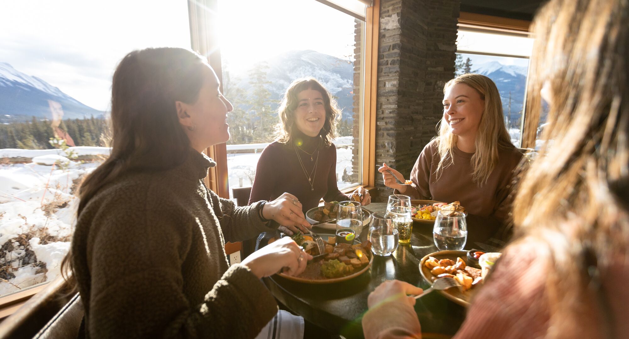 Four women sit at a table at Juniper Bistro in Banff enjoying a cozy brunch in the winter with mountain views out the window.