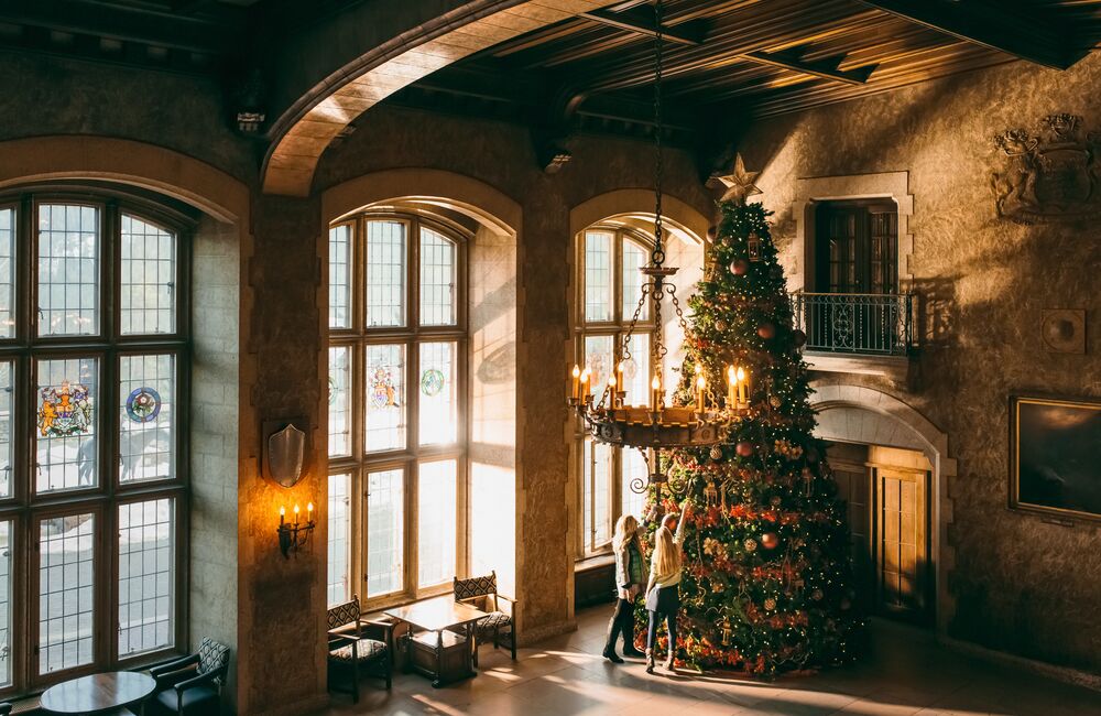 Two women look at a massive Christmas tree inside a grand hall at the Banff Springs Hotel. There are three windows along the wall illuminating the tree in Banff National Park.