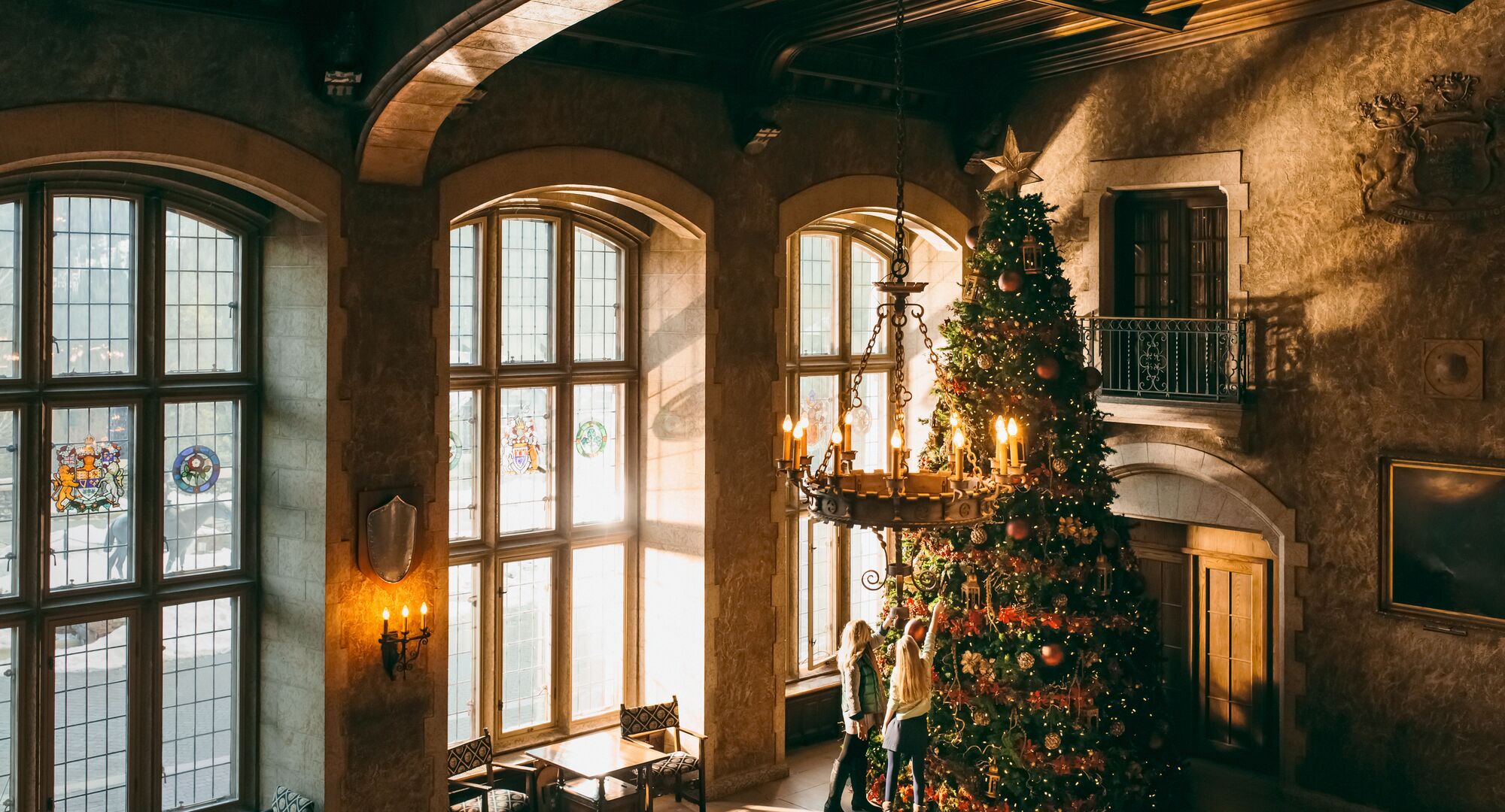 Two women look at a massive Christmas tree inside a grand hall at the Banff Springs Hotel. There are three windows along the wall illuminating the tree in Banff National Park.
