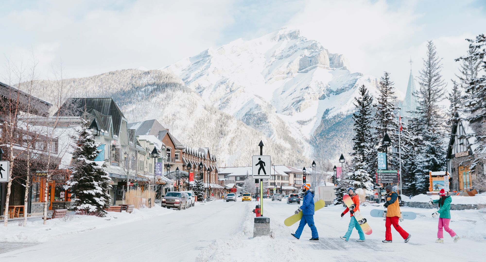 Four snowboarders cross Banff Avenue with Cascade Mountain in the background.