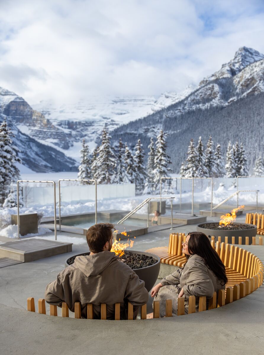 Two people sit at a fire pit outside at the BASIN spa at the Chateau Lake Louise in the winter.