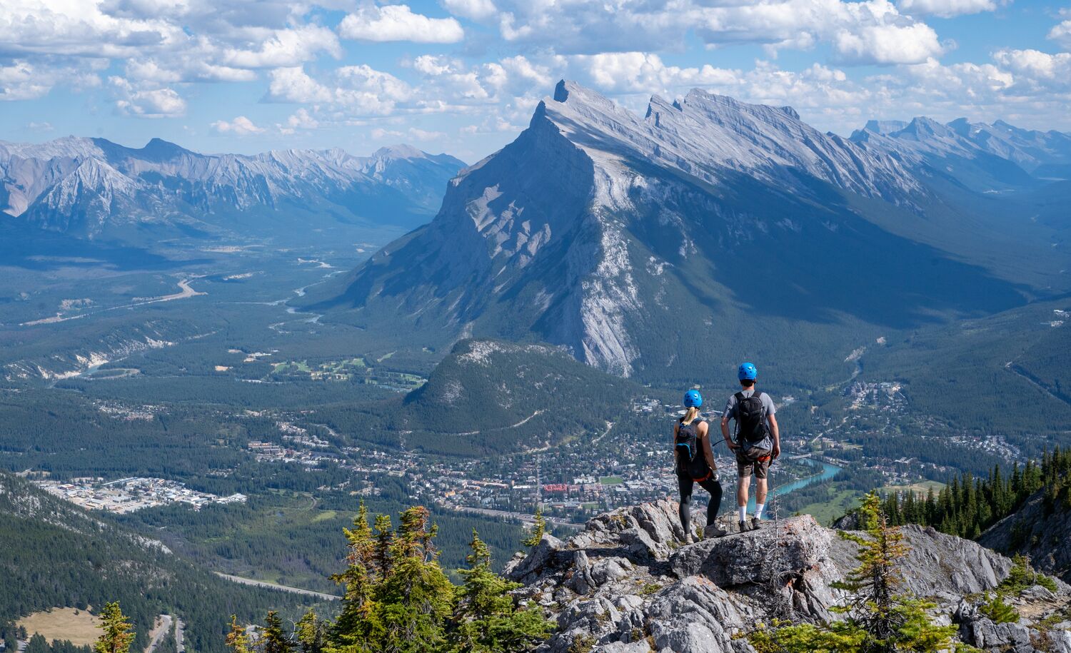 Two people lookout at the Banff townsite from the Mt. Norquay via ferrata on a bluebird day with views of Mount Rundle.