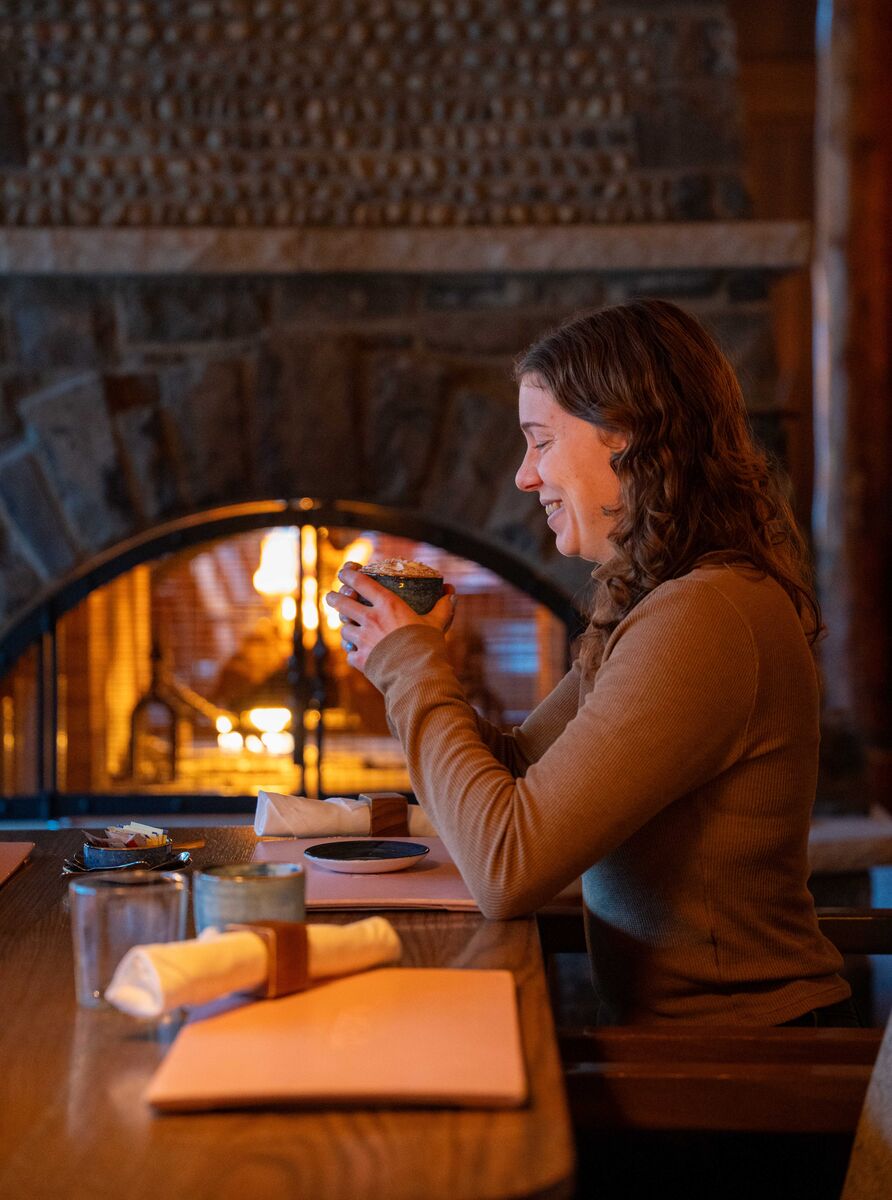 A woman enjoys a hot chocolate in front of a fireplace at Bluebird in Banff.